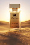 A rectangular glass perfume bottle labeled Karaama Oud de Amber stands centered on golden sand dunes at sunset, with a soft blurred desert landscape behind it.